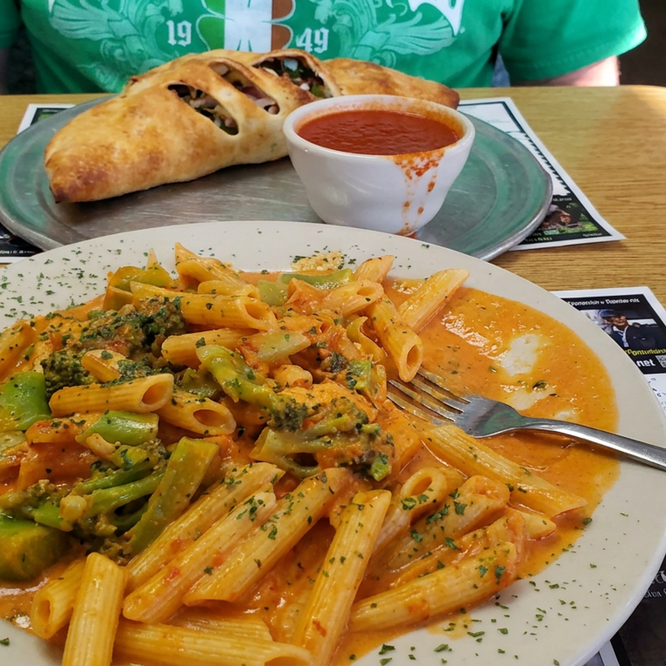 Penne pasta with broccoli in creamy tomato sauce, served alongside a calzone and marinara dipping cup at a restaurant table.