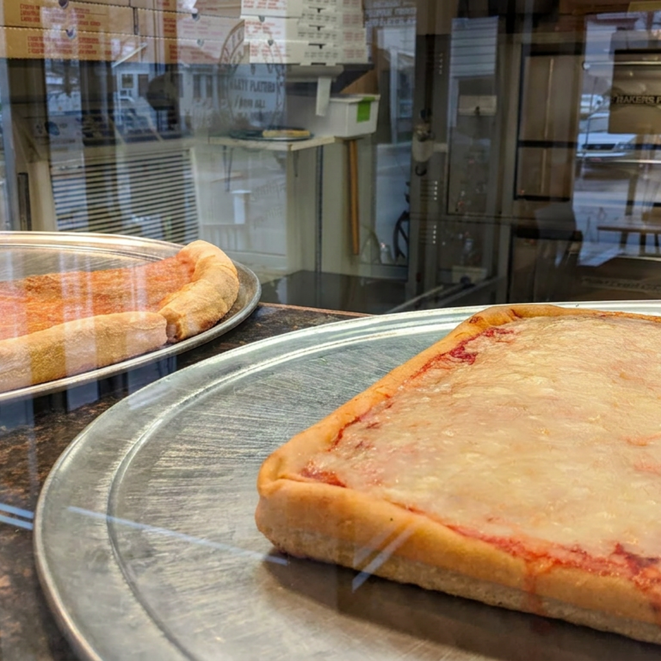 Two rectangular cheese pizzas sit on metal serving trays behind a restaurant display window.